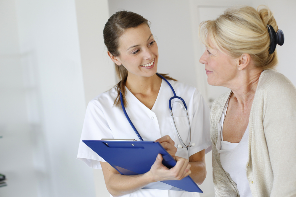 Clinician with stethoscope and clipboard smiling at blonde elderly patient